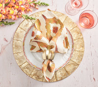 An elegant table is set with Kim Seybert Capiz Shell Placemat Champagne, gold-rimmed and white plates, a patterned napkin in a gold ring, two pink drinks, and orange flowers on a light pink marble surface.