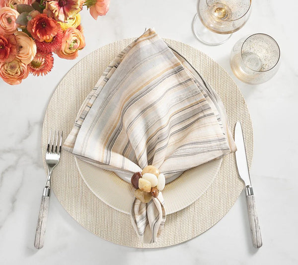 A table setting features a striped napkin in a Kim Seybert Sea Stone Napkin Ring Beige Set of 4 on a white plate, beside cutlery, two drinks, and orange flowers on a white surface.