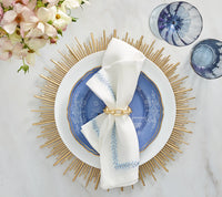 Create a stunning table with Kim Seybert's Jardin Napkin Set of 4, featuring blue embroidery in gold rings, paired with a blue patterned plate, sunburst placemat, blue glasses, and pink flowers atop a marble surface.