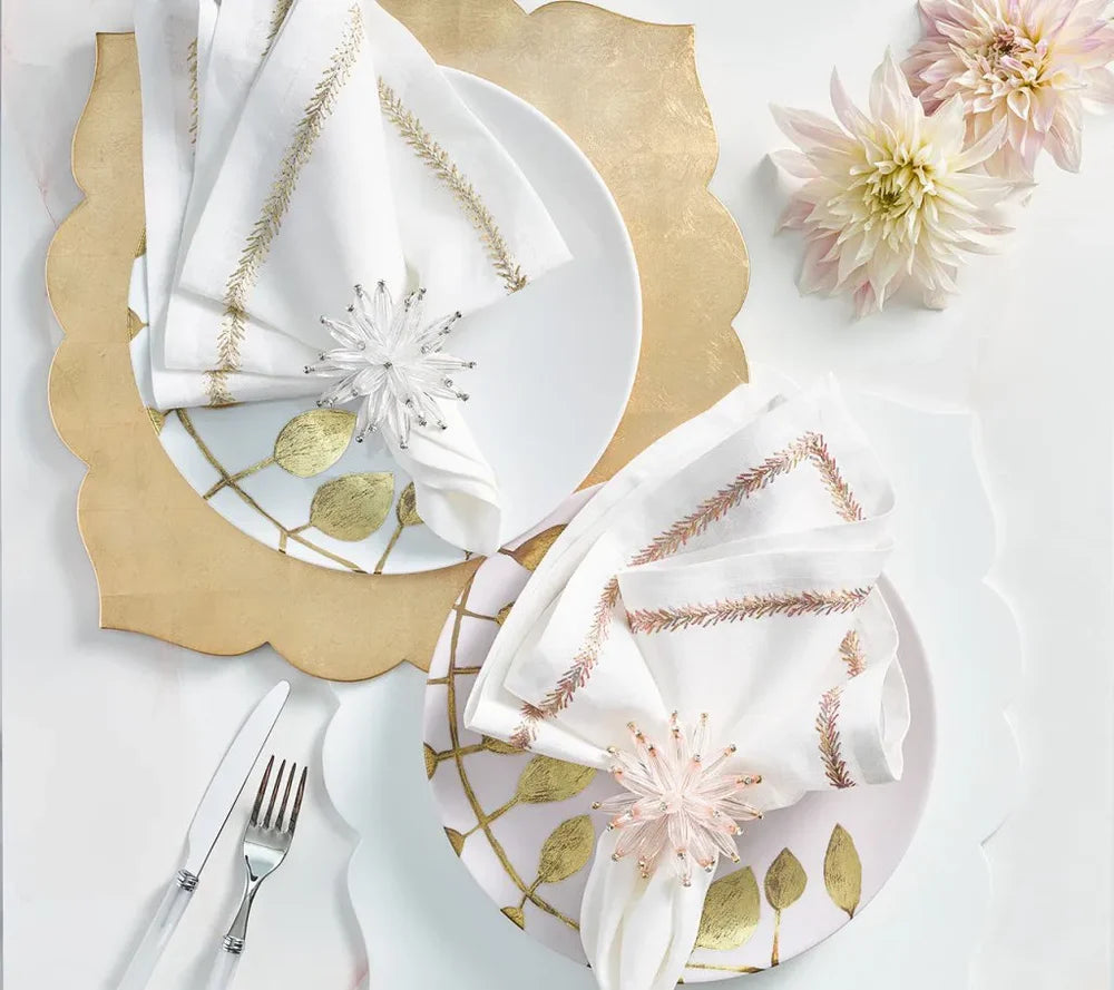 Two elegant place settings featuring Guzzini’s Fez Placemats in a chic champagne scallop design, white plates adorned with gold leaf patterns, white napkins embroidered with gold, clear and pink napkin rings, and two dahlias in the top right corner. The fork and knife are placed to the left of each setting.