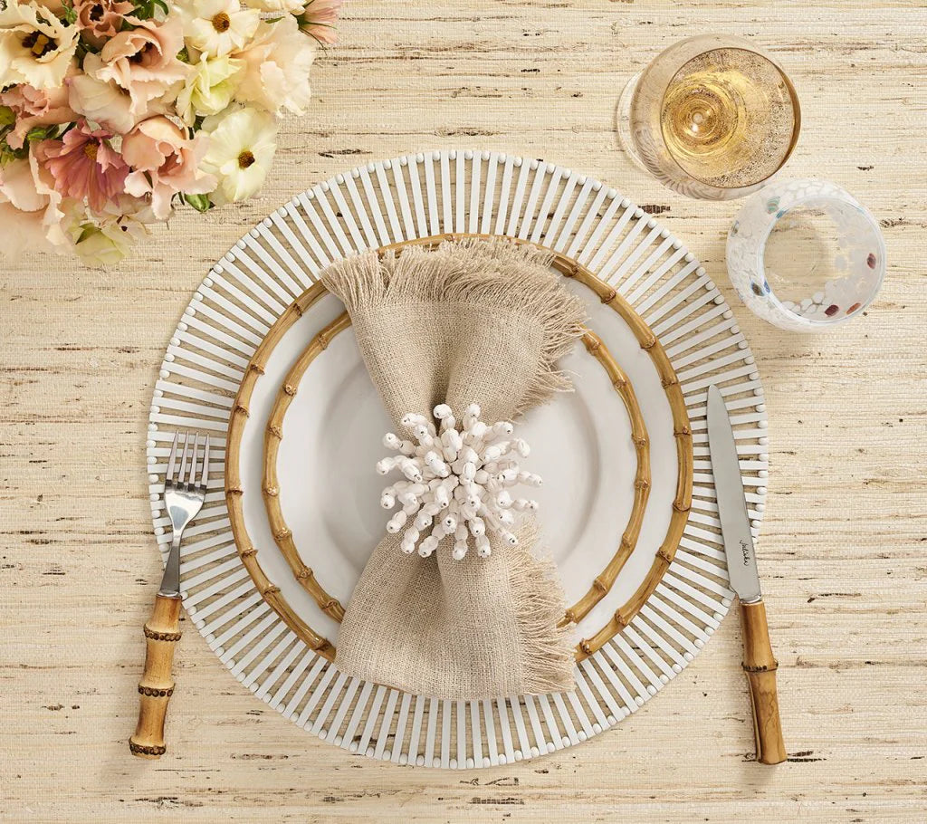 A place setting features a white and bamboo-patterned plate, Kim Seybert Fringe Napkin Silver & Natural, bamboo-handled cutlery, a wine glass, water glass, and a floral centerpiece on a textured table.
