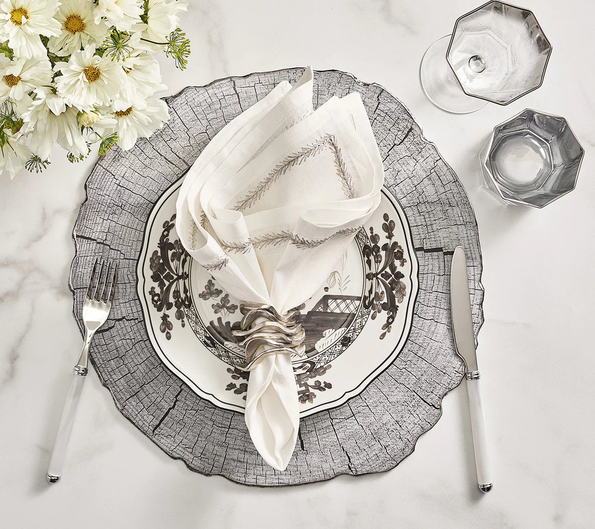 The Jardin Napkin Set of 4 by Kim Seybert is styled with a floral plate, silver-ringed napkin, white-handled fork and knife, two glasses, and a flower bouquet on a wood-patterned placemat atop marble.