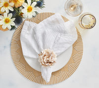 A white napkin with a beige flower ring sits on a white plate atop a Kim Seybert Dream Weaver Placemat Natural, with glasses of sparkling drink and water nearby and a bouquet of white and yellow flowers.