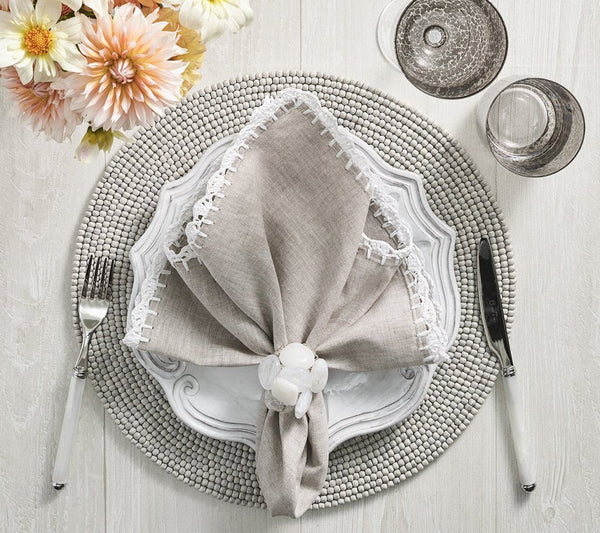 Create an elegant table setting with the Kim Seybert Driftwood Placemat Set 4 Grey, paired with white plates, a beige lace-trimmed napkin and ring, silver cutlery, gray glasses, and dahlias—perfect handcrafted décor for any occasion.