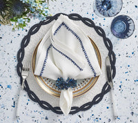 The Filament Napkin White & Navy Set of 4 by Kim Seybert adds elegance to your table with neatly folded white napkins featuring blue trim, paired with silverware, navy napkin rings, blue glasses, and a floral centerpiece on a speckled surface.