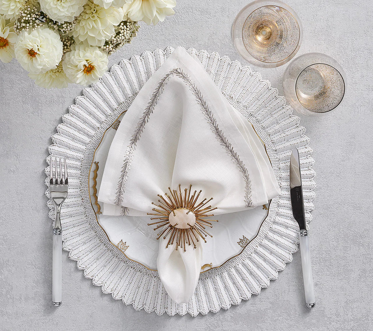 An elegant white napkin is adorned with a Kim Seybert Flare Napkin Ring Gold Set of 4, displayed on a white placemat. A bouquet of white dahlias appears at the top left.