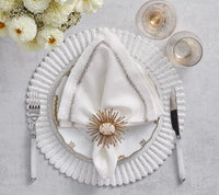 An elegant white napkin is adorned with a Kim Seybert Flare Napkin Ring Gold Set of 4, displayed on a white placemat. A bouquet of white dahlias appears at the top left.