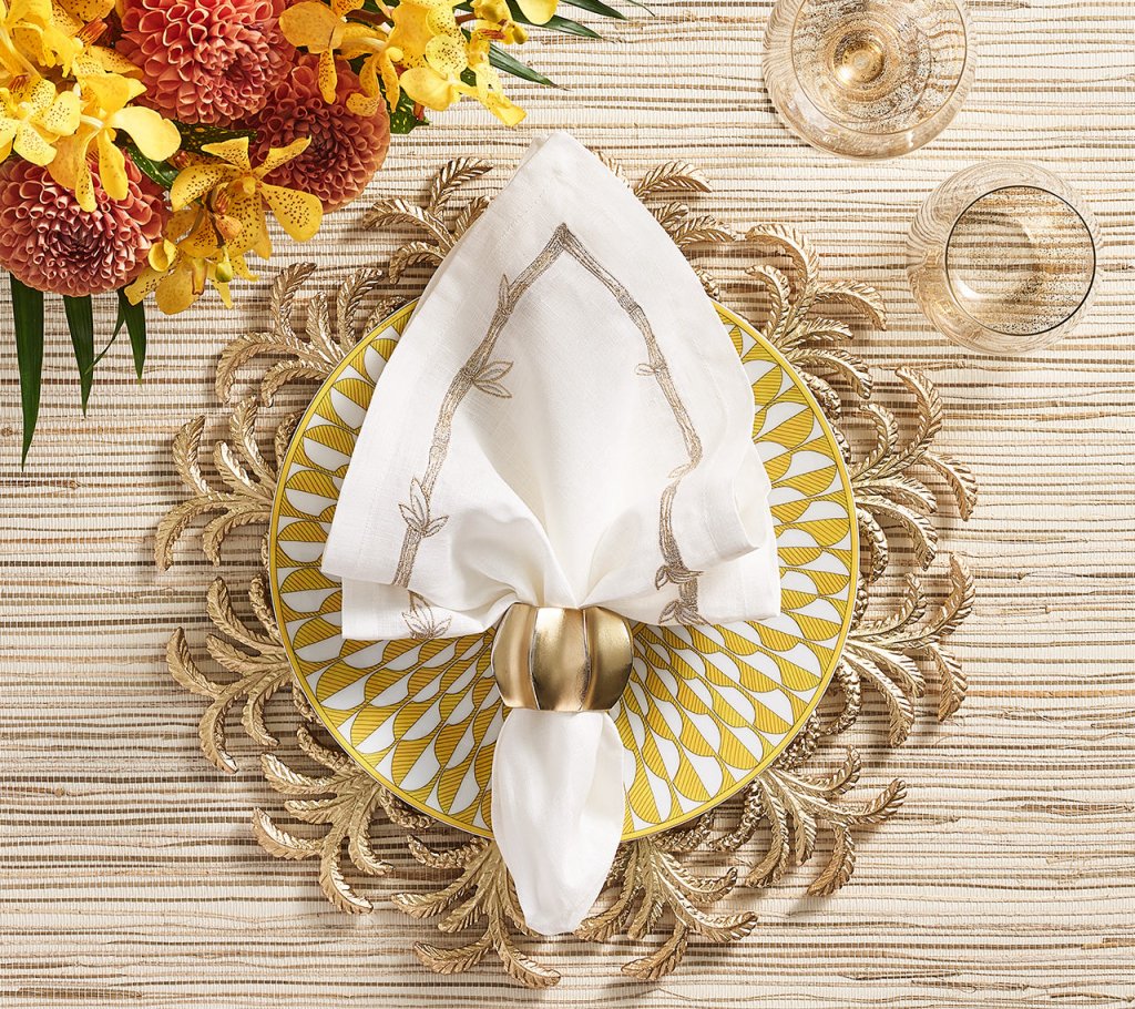 A table setting features a yellow and white patterned plate on a gold leaf placemat, topped with a folded white napkin in a Kim Seybert Desert Napkin Ring Gold & Silver (set of 4). Nearby are two glasses and a yellow-orange flower arrangement.