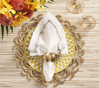 A table setting features a yellow and white patterned plate on a gold leaf placemat, topped with a folded white napkin in a Kim Seybert Desert Napkin Ring Gold & Silver (set of 4). Nearby are two glasses and a yellow-orange flower arrangement.