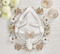 An elegant table setting features white florals, gold accents, a scalloped-edge plate, and a white napkin with a Kim Seybert Papillon Butterfly Napkin Ring (Boxed Set of 4), plus two glasses on a textured white surface—perfect for luxury decor.