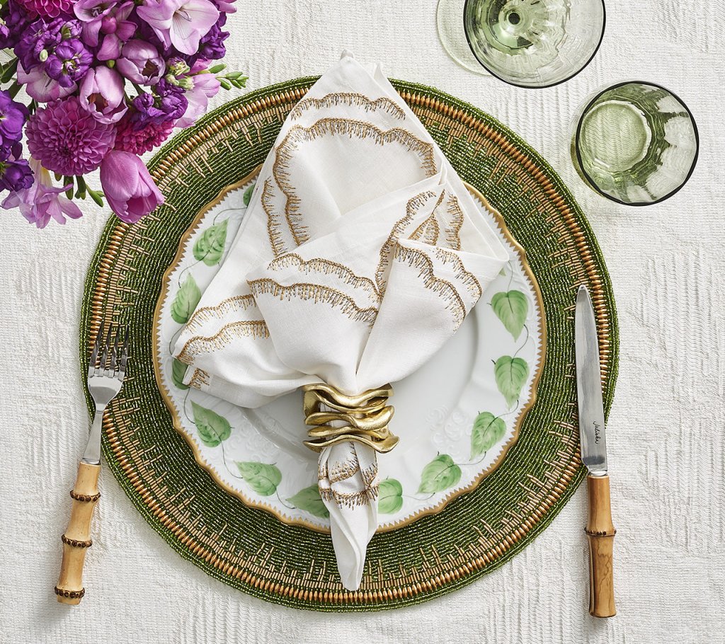An elegant table setting featuring Kim Seybert’s Flux Napkin Ring Gold Set of 4 on white napkins, with a floral plate, green charger, bamboo-handled flatware, two green glasses, and a purple flower arrangement on a white textured tablecloth.
