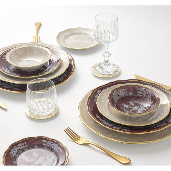Elegant table setting with Ginori 1735 Oriente Italiano Beige Meringa porcelain dinnerware, gold-trimmed and stacked in burgundy and cream, paired with gold utensils and clear glassware on a white surface.