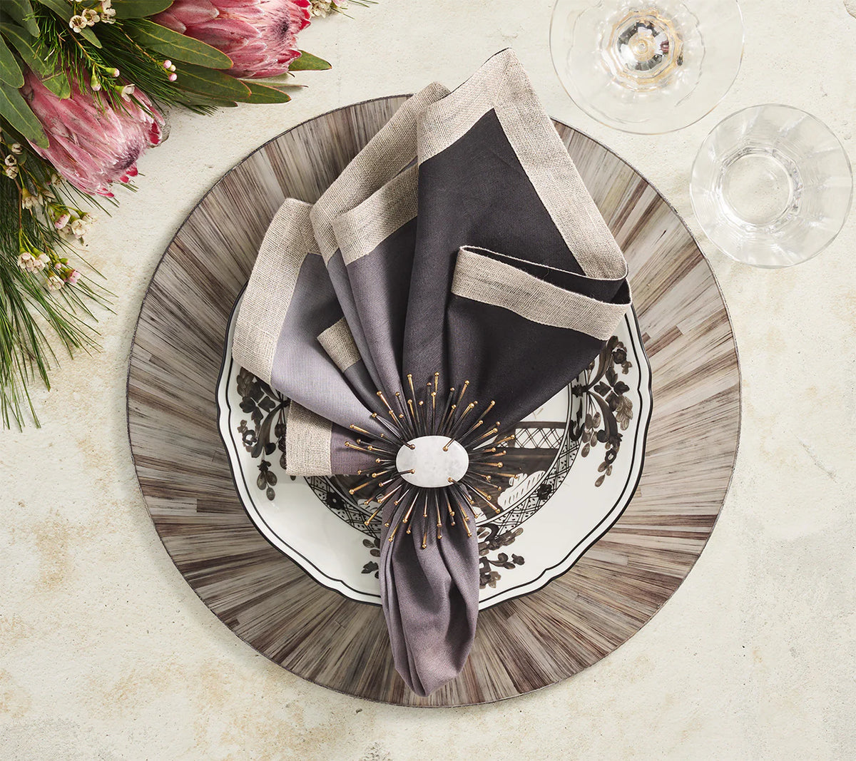 A decorative place setting features a patterned black and white plate on a gray charger, topped with a folded napkin in an ornate ring. Wine glasses and flowers sit atop Kim Seybert’s Aura Placemat Grey Set of 4, adding a high-gloss finish to the elegant look.