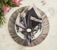 A decorative place setting features a patterned black and white plate on a gray charger, topped with a folded napkin in an ornate ring. Wine glasses and flowers sit atop Kim Seybert’s Aura Placemat Grey Set of 4, adding a high-gloss finish to the elegant look.