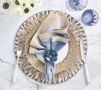 A decorative table setting featuring a beaded placemat, white plate, blue and beige napkin accented with a Kim Seybert Begonia Napkin Ring in teal (set of 4), silver fork and knife, and two blue glasses on a marble surface.