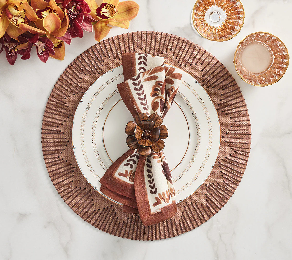 A white dinner plate with gold trim sits on a Kim Seybert Dream Weaver Placemat Set of 4 in brown. A brown floral napkin with leaf pattern is held by a wooden flower napkin ring. Orange and burgundy orchids rest to the left.