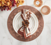 A white dinner plate with gold trim sits on a Kim Seybert Dream Weaver Placemat Set of 4 in brown. A brown floral napkin with leaf pattern is held by a wooden flower napkin ring. Orange and burgundy orchids rest to the left.