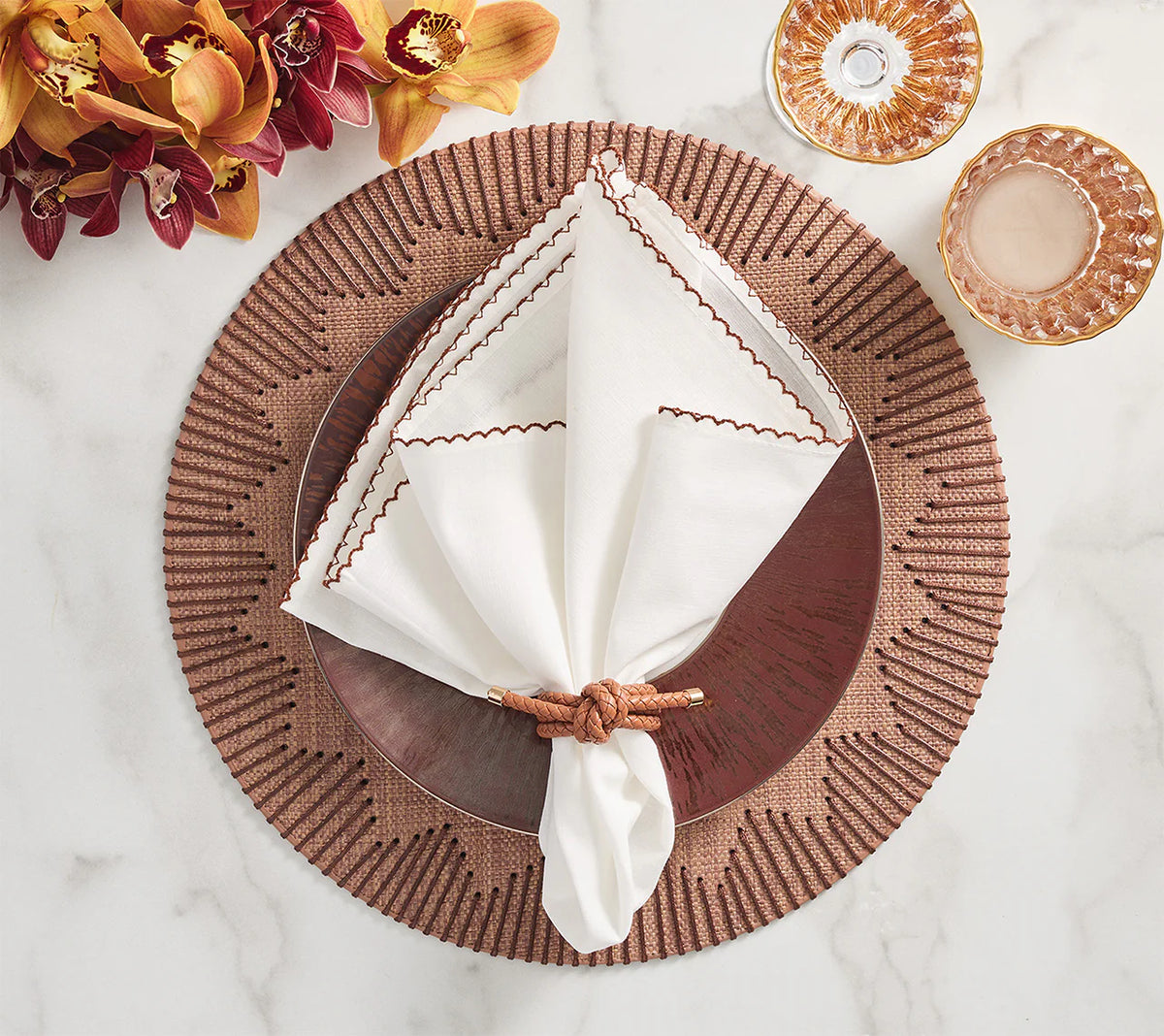 A table setting with a set of 4 Kim Seybert Dream Weaver brown placemats, a dark brown plate, white napkin with brown trim in a braided ring, two pink glass cups, and yellow and burgundy flowers.