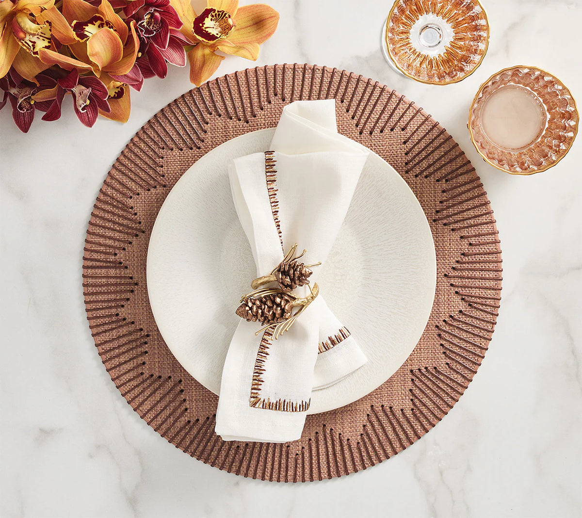 A table is styled with a white plate on Kim Seybert’s Dream Weaver Placemat Set of 4 in brown, a white napkin with decorative stitching and pinecone ring, two pink glasses, and orange orchids displayed on a marble surface.