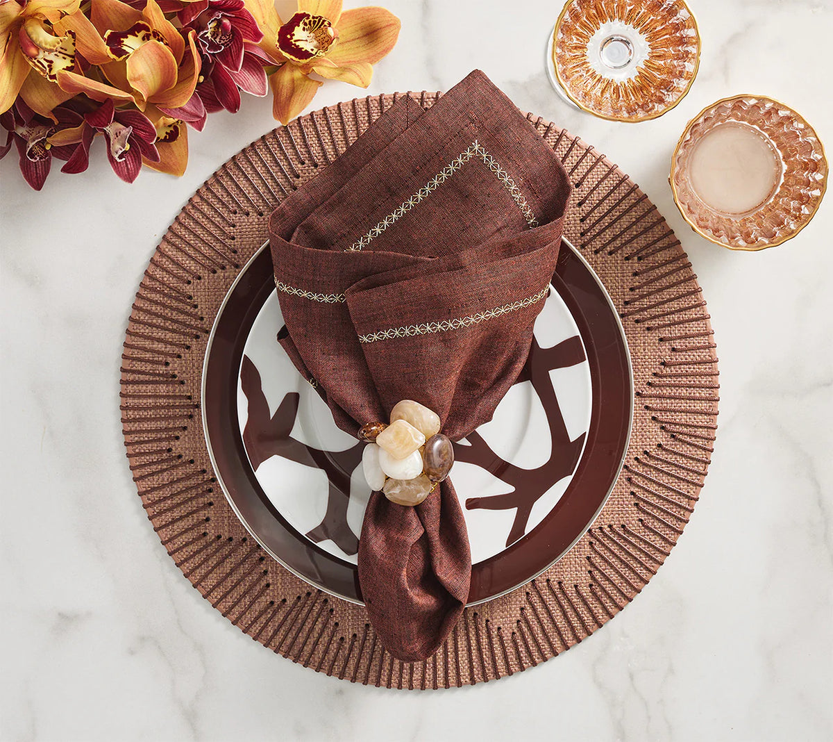 A decorative table setting features a maroon napkin in a jeweled ring on a patterned plate, brown Kim Seybert Dream Weaver Placemats (Set of 4), gold-rimmed glassware, and a bouquet of burgundy and yellow flowers on marble.