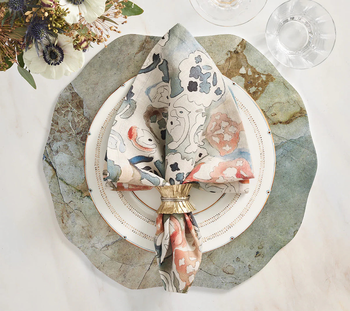 A decorative place setting features a scalloped stone placemat, white plate with gold rim, linen napkin from the Kim Seybert Giverny Napkin Set of 4 - Multi in a gold ring, clear glassware on marble, and a Monet-inspired floral arrangement.
