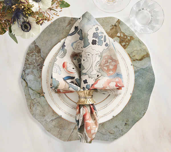 A decorative place setting features a scalloped stone placemat, white plate with gold rim, linen napkin from the Kim Seybert Giverny Napkin Set of 4 - Multi in a gold ring, clear glassware on marble, and a Monet-inspired floral arrangement.