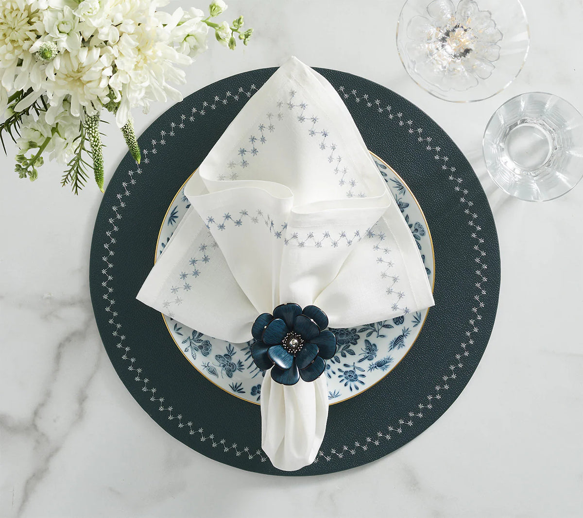 A blue-gray placemat with a patterned plate, a white napkin adorned with blue stars and accented by Kim Seybert’s Begonia Napkin Rings Set of 4 in Teal, a glass of water, a glass bowl, and white flowers arranged on a marble table.