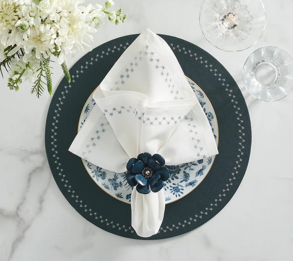 A blue-gray placemat with a patterned plate, a white napkin adorned with blue stars and accented by Kim Seybert’s Begonia Napkin Rings Set of 4 in Teal, a glass of water, a glass bowl, and white flowers arranged on a marble table.