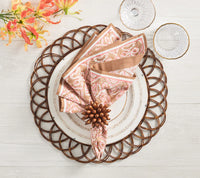 A table setting featuring Kim Seybert's Florette Napkin Ring Wood Set of 4 with a woven charger, white plate, pink and brown patterned napkin, crystal glasses, and orange flowers adds rustic charm to a light wood table.