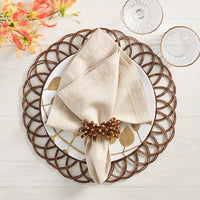 A beige napkin with the Kim Seybert Royal Palm Napkin Ring Set of 4 rests on a white plate with gold leaf accents atop a woven brown placemat, surrounded by two clear glass dishes and orange flowers on a light wood table for tropical flair.