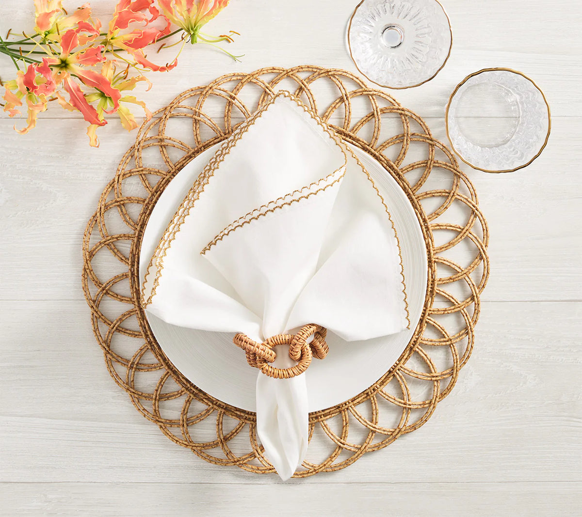 A white plate rests on the Kim Seybert Alfresco Placemat Set of 4 in Natural, topped with a gold-trimmed napkin and ring. Two clear glasses and orange flowers adorn this inviting outdoor table set on light wood.
