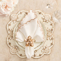 A scalloped, embroidered placemat with seashell motifs holds stacked white plates and a gold-trimmed napkin secured by the Kim Seybert Beachcomber Napkin Ring Set of 4. Glassware and pale flowers bring coastal elegance to the marble surface.