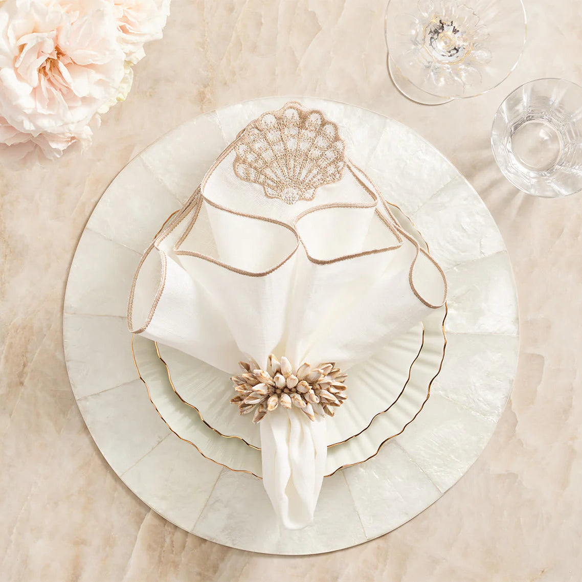 An elegant beach-inspired table setting features a white plate, scalloped salad plate, a gold-embroidered napkin with a Kim Seybert Shell Fringe Napkin Ring Set of 4, crystal glasses, and pale pink flowers atop a marble surface.