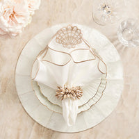 An elegant beach-inspired table setting features a white plate, scalloped salad plate, a gold-embroidered napkin with a Kim Seybert Shell Fringe Napkin Ring Set of 4, crystal glasses, and pale pink flowers atop a marble surface.