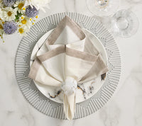 A Kim Seybert Dip Dye Napkin in earthtones is folded atop a white plate with black marble patterns on a silver charger. The napkin, secured by a clear ring, is set beside wine glasses and a floral arrangement.