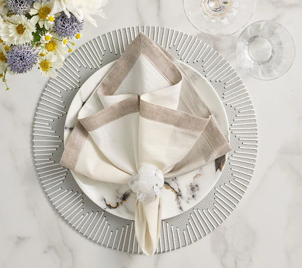 A Kim Seybert Dip Dye Napkin in earthtones is folded atop a white plate with black marble patterns on a silver charger. The napkin, secured by a clear ring, is set beside wine glasses and a floral arrangement.