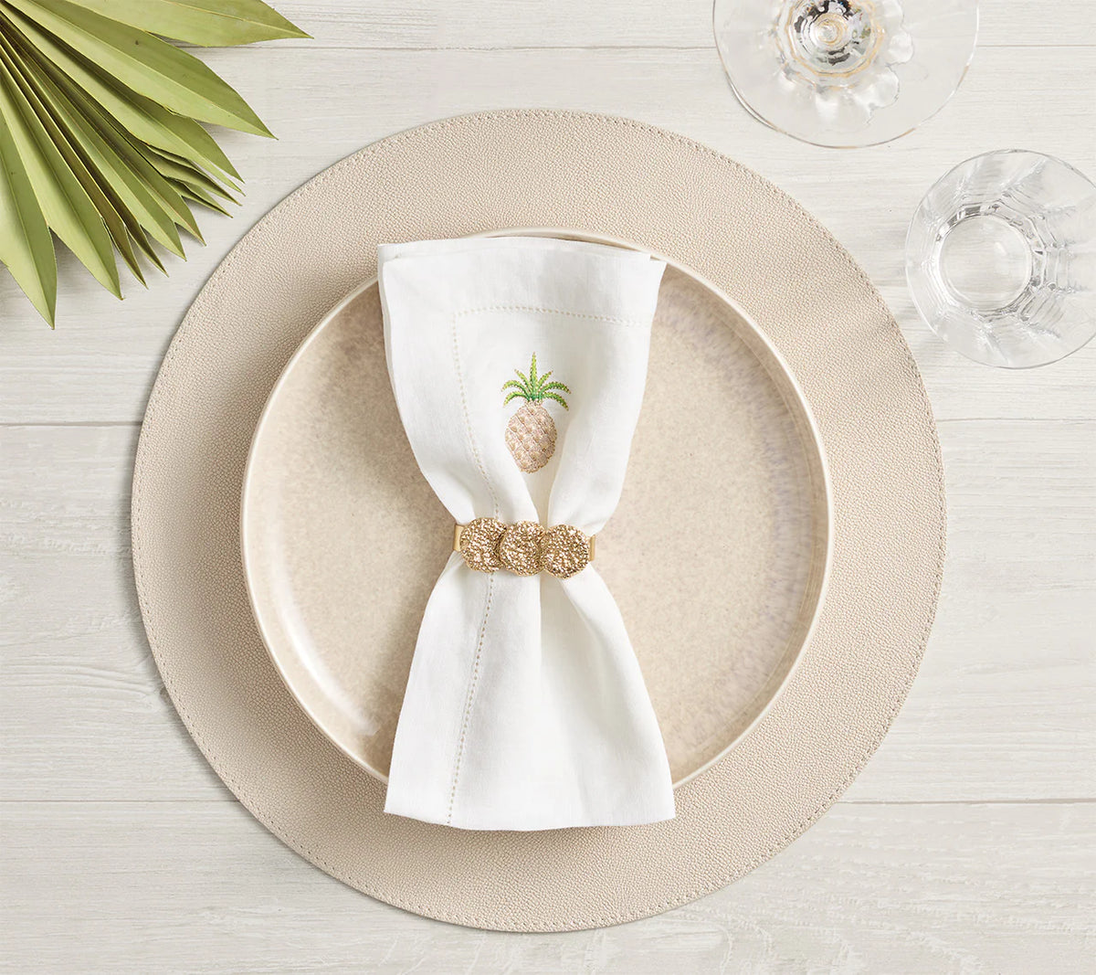 A beige dinner plate rests on a Kim Seybert Icons Placemat from the set of 4. A white napkin with a gold pineapple and gold napkin ring, plus wine and water glasses, decorate the light wood table with green leaves nearby.