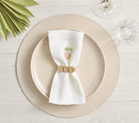 A beige dinner plate rests on a Kim Seybert Icons Placemat from the set of 4. A white napkin with a gold pineapple and gold napkin ring, plus wine and water glasses, decorate the light wood table with green leaves nearby.