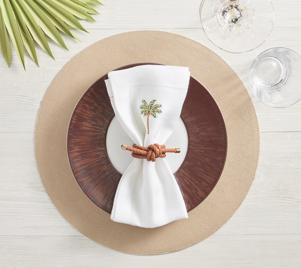 A table is set with a Kim Seybert Icons Placemat, dark brown and white plates, a white napkin with an embroidered palm tree in a rope ring, plus wine and water glasses and a palm leaf nearby.