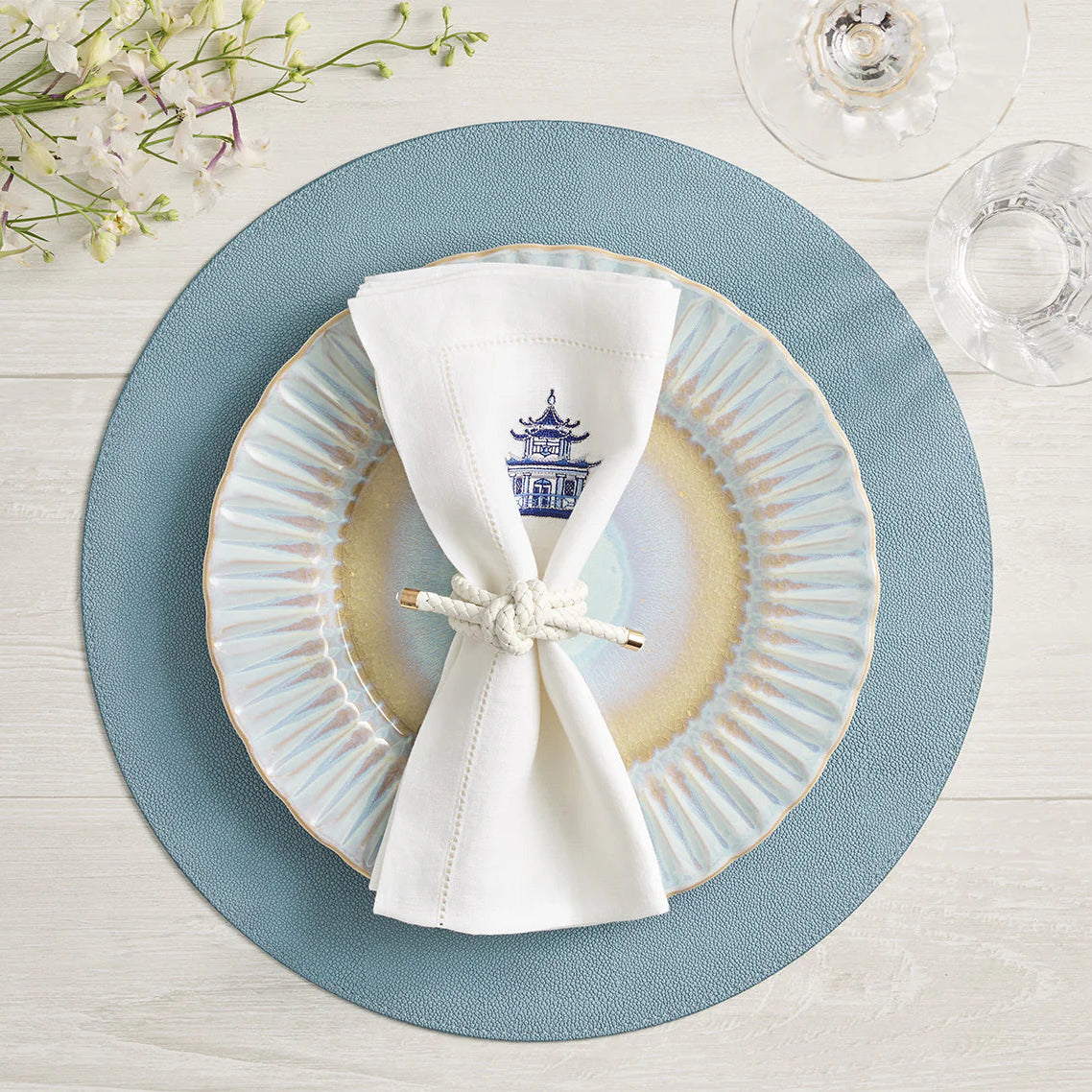 A blue and beige ceramic plate rests on a Kim Seybert Icons Placemat, paired with a white napkin featuring a blue pagoda design, neatly knotted. A wine glass and flowers complete the setting on the light wood table.