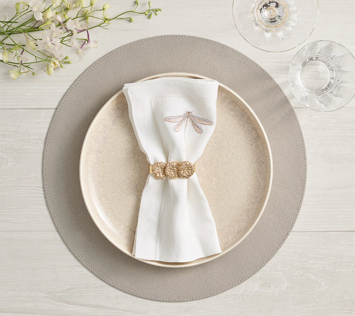 A beige plate rests on a Kim Seybert Icons Placemat with subtle shagreen texture, topped with a dragonfly-embroidered napkin and gold ring. A water glass, wine glass, and white flowers sit nearby on the light wood table.