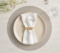 A beige plate rests on a Kim Seybert Icons Placemat with subtle shagreen texture, topped with a dragonfly-embroidered napkin and gold ring. A water glass, wine glass, and white flowers sit nearby on the light wood table.