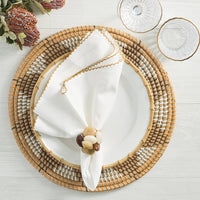 A white plate sits on a woven placemat with a white napkin folded through a Kim Seybert Sea Stone Napkin Ring Beige (Set of 4). Two clear glass dishes and a floral arrangement rest nearby on the light wood table.