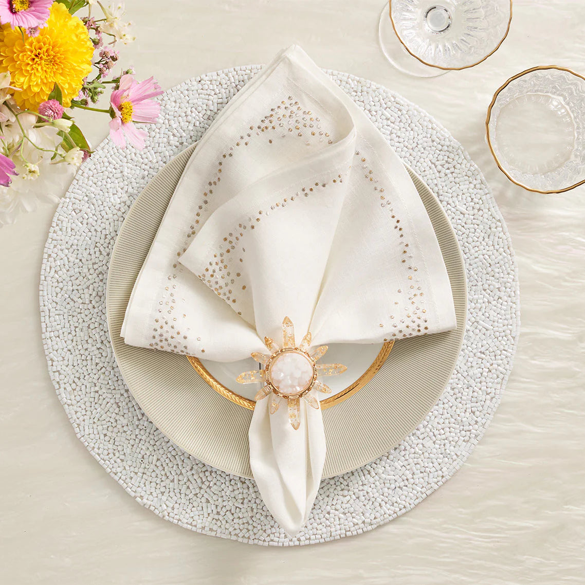 An elegant table setting featuring the Kim Seybert Pin Dot Napkin Set of 4, stylishly folded on a white plate, complemented by a gold-rimmed glass, a flower-shaped napkin ring, a textured placemat, and a small bouquet of pink and yellow flowers in the corner.