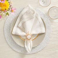 An elegant table setting featuring the Kim Seybert Pin Dot Napkin Set of 4, stylishly folded on a white plate, complemented by a gold-rimmed glass, a flower-shaped napkin ring, a textured placemat, and a small bouquet of pink and yellow flowers in the corner.
