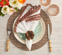 A rustic table features the Kim Seybert Shell Edge Napkin (set of 4) with brown trim, folded in a wooden flower ring on a green striped plate. Bamboo-handled utensils and a bouquet of orange and white flowers complete the look.