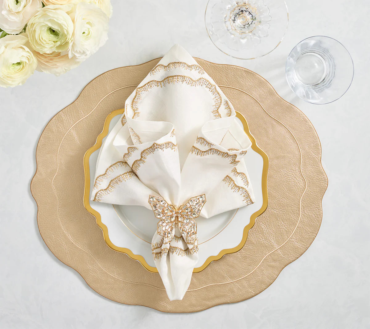 Create a luxurious table setting with layered beige and white plates, an embroidered napkin held by a Kim Seybert Papillon Butterfly Napkin Ring, clear glasses on a white tablecloth, and a bouquet of white flowers.