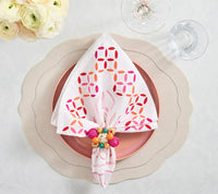 A white napkin with a pink and orange geometric pattern sits in a beaded ring on a pink plate and scalloped cream charger, all atop the Kim Seybert Tailored Placemat Set of 4 in Ivory, beside a glass and fresh white flowers.