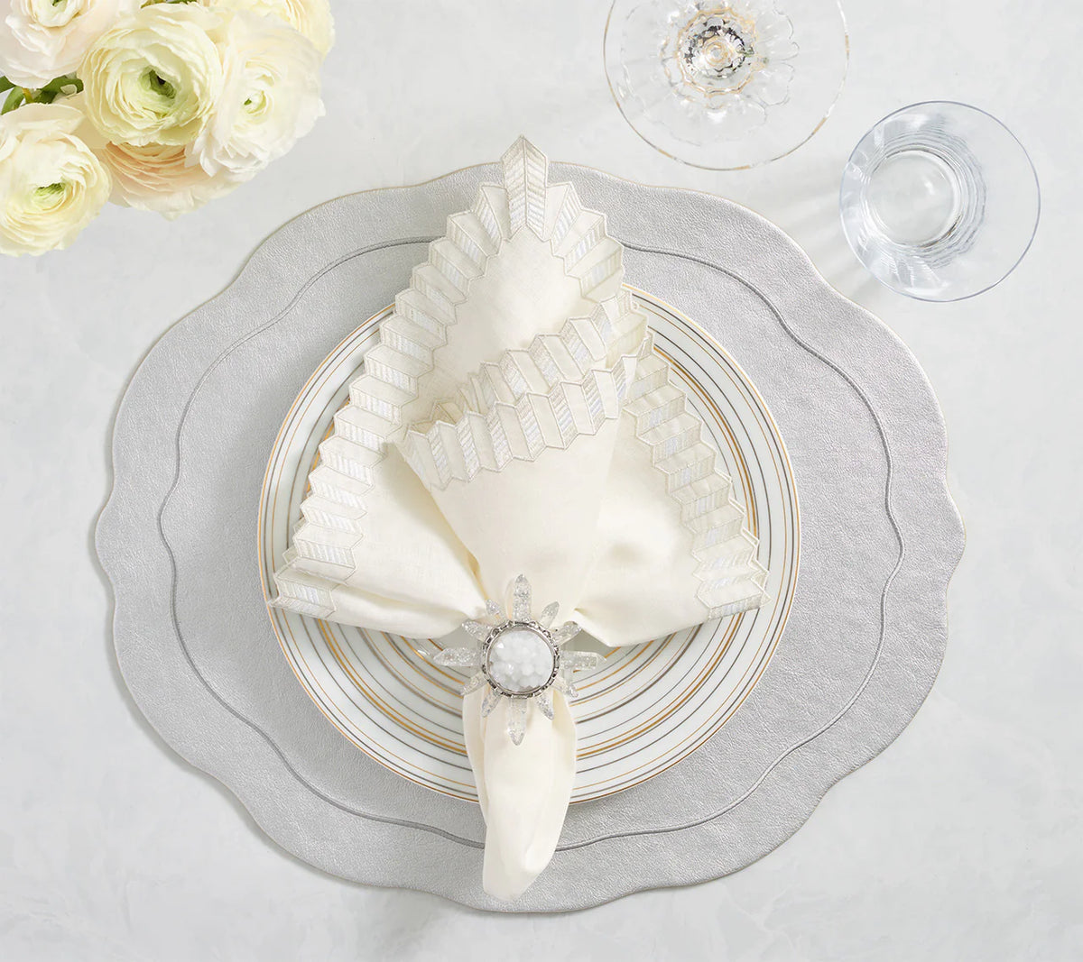 An elegant table setting with a cream and silver scalloped napkin in a jeweled holder, set on layered plates atop the Kim Seybert Tailored Placemat Set of 4 - Silver, beside clear glassware and white flowers on light marble.