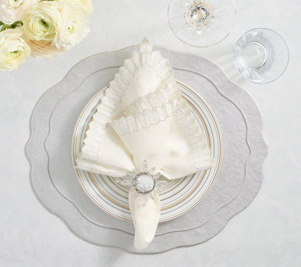 An elegant table setting with a cream and silver scalloped napkin in a jeweled holder, set on layered plates atop the Kim Seybert Tailored Placemat Set of 4 - Silver, beside clear glassware and white flowers on light marble.